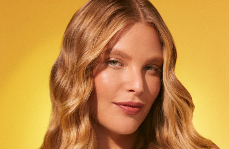 Foto com fundo amarelo. Mulher com cabelo ondulado e comprido, loiro, usando blusa branca.